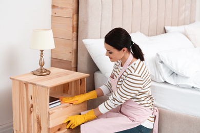 Photo of Young chambermaid wiping dust from nightstand in bedroom