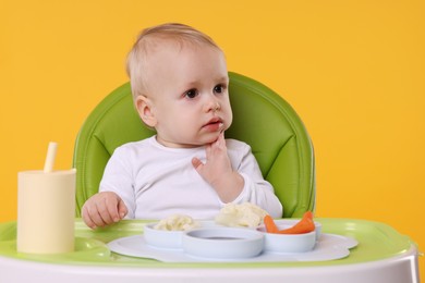 Cute little baby with healthy food in high chair on orange background