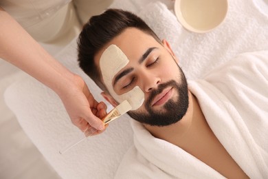 Photo of Cosmetologist applying mask on man's face in spa salon, top view