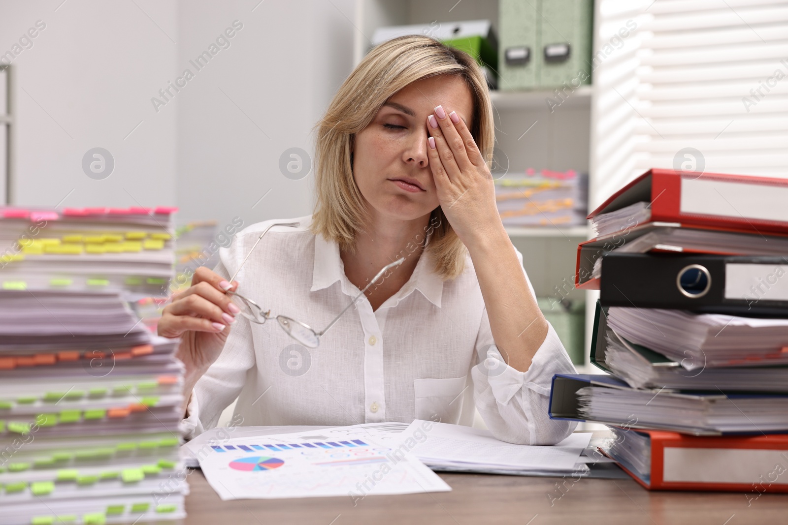 Photo of Overwhelmed woman sitting at table with stacks of documents and folders in office