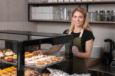 Happy seller taking delicious dessert from showcase in bakery shop