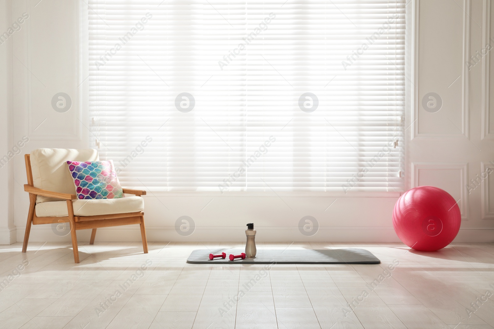 Photo of Exercise mat, dumbbells, bottle and fitness ball near armchair in spacious room