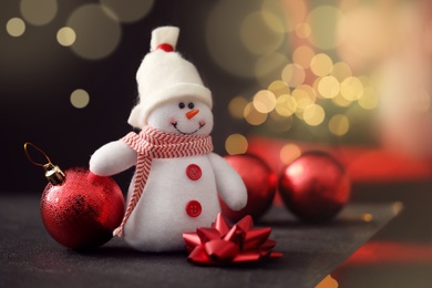 Photo of Closeup view of snowman toy, Christmas ball and bow on black table against blurred festive lights. Space for text