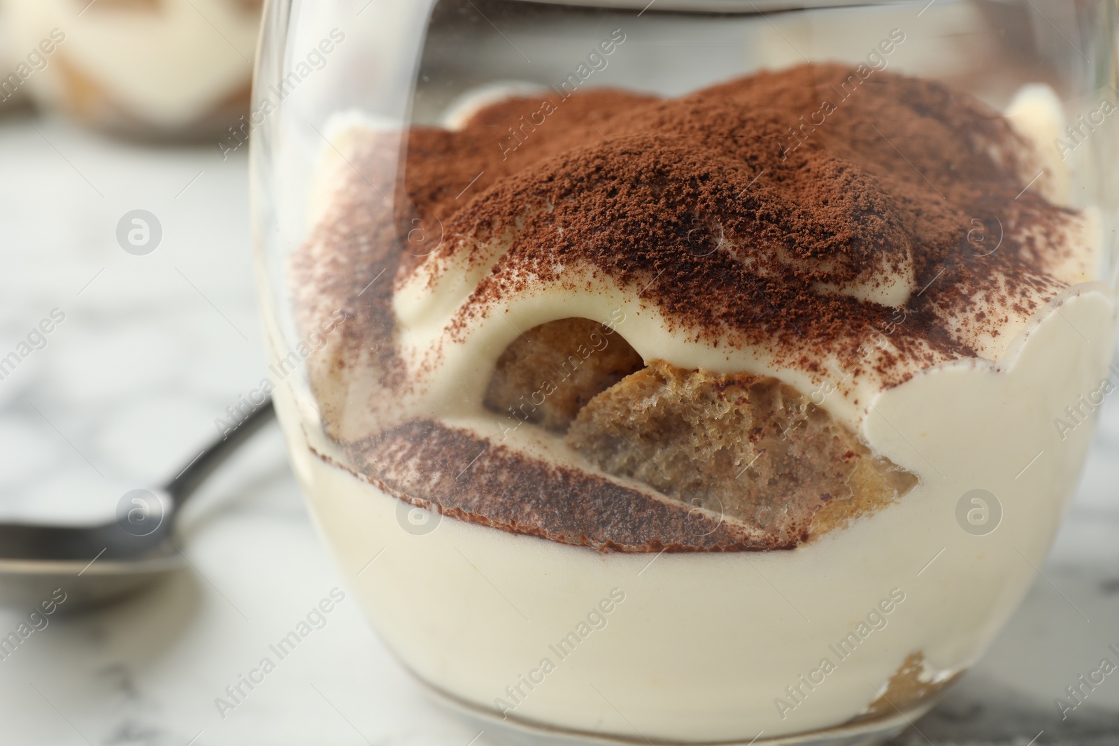 Photo of Delicious tiramisu in glass and spoon on white marble table, closeup