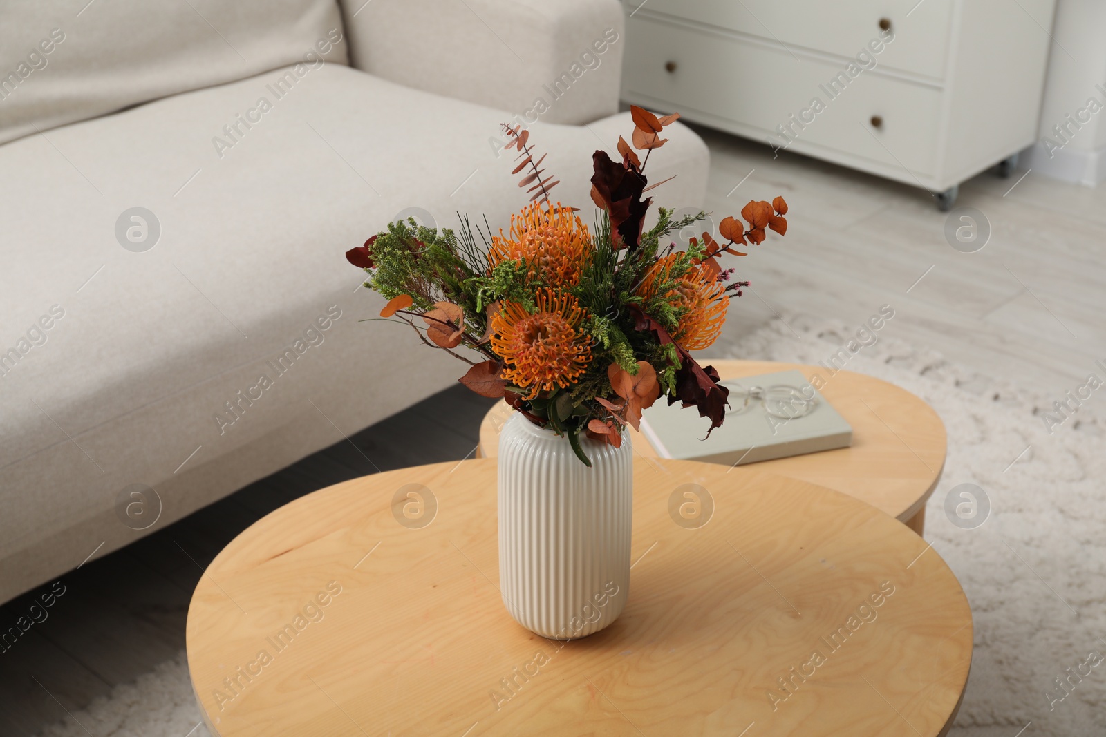 Photo of Vase with bouquet of beautiful leucospermum flowers, book and glasses on wooden nesting tables indoors