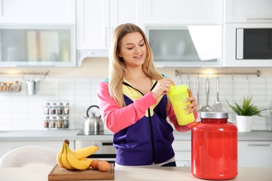 Young woman holding bottle of protein shake near table with ingredients in kitchen