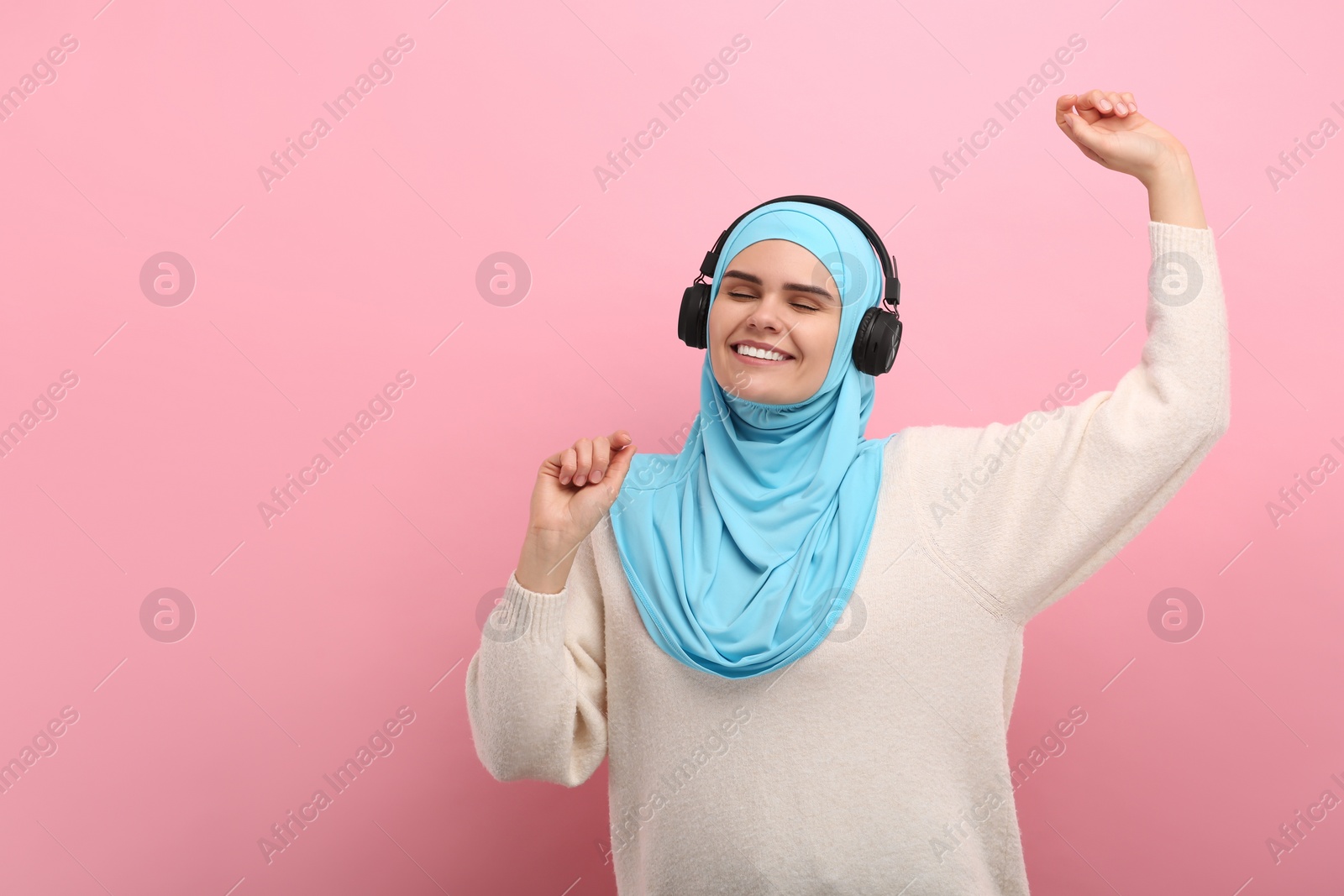 Photo of Muslim woman in hijab and headphones listening to music on pink background, space for text
