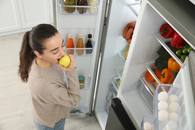 Photo of Young woman eating apple near open refrigerator indoors, above view