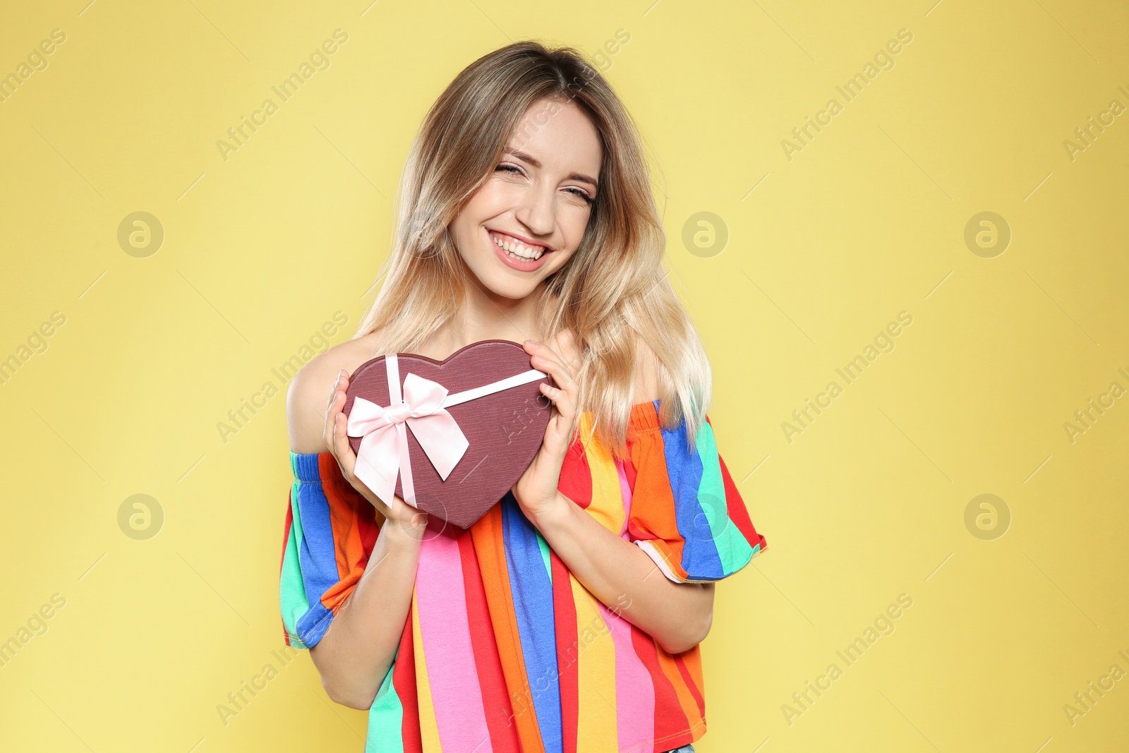 Photo of Portrait of beautiful smiling girl with heart shaped gift box on yellow background. International Women's Day