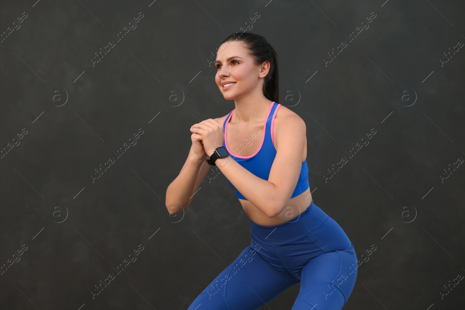 Photo of Beautiful woman in stylish sportswear doing squats near black wall