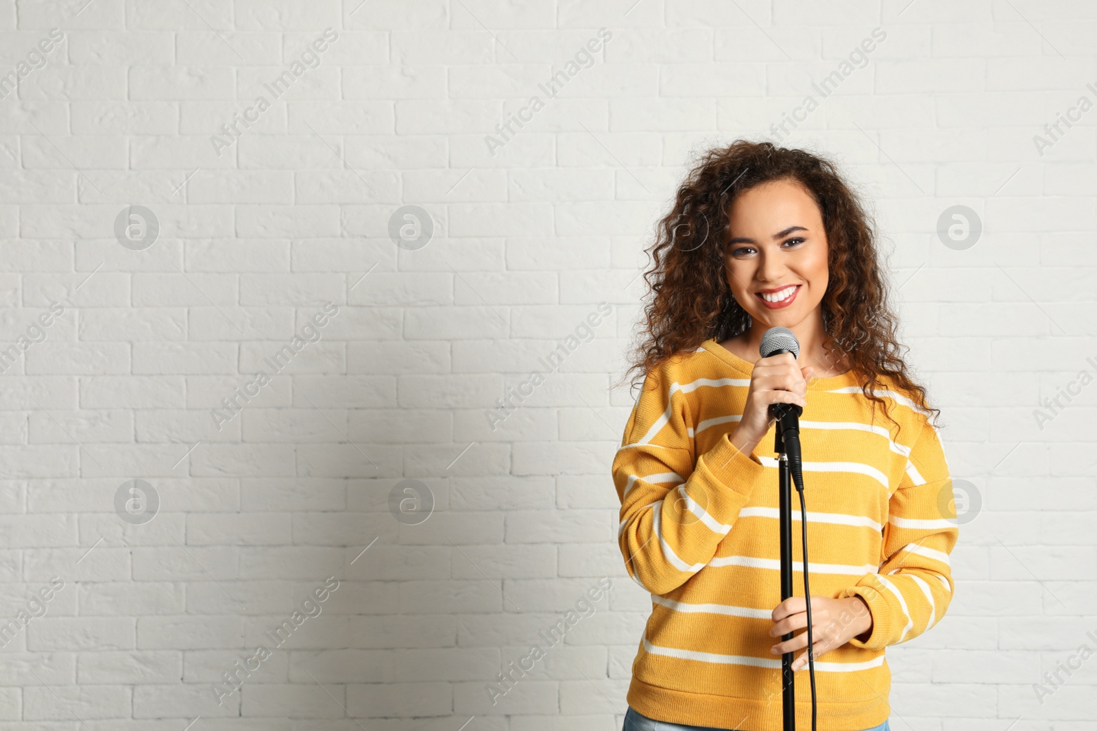 Photo of Portrait of curly African-American woman in sweater posing with microphone near brick wall. Space for text