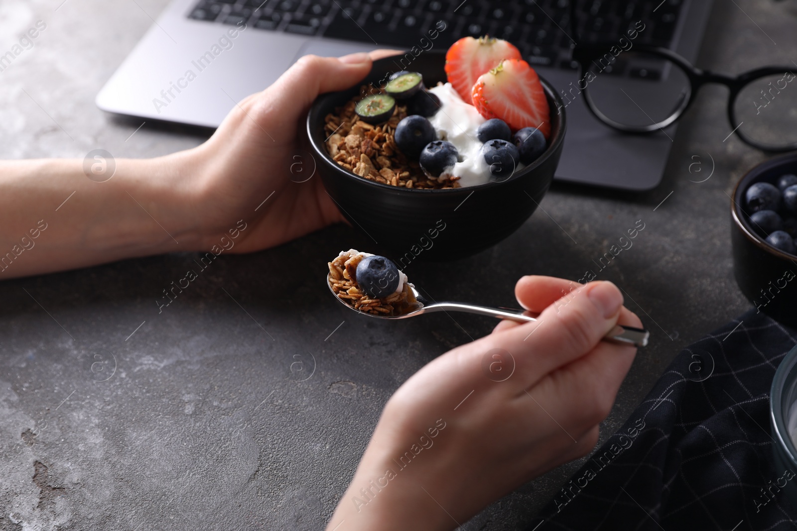 Photo of Woman eating tasty granola with yogurt and berries at workplace, closeup