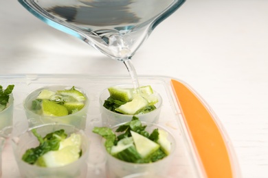 Pouring water into ice cube tray with lime slices and mint on table, closeup