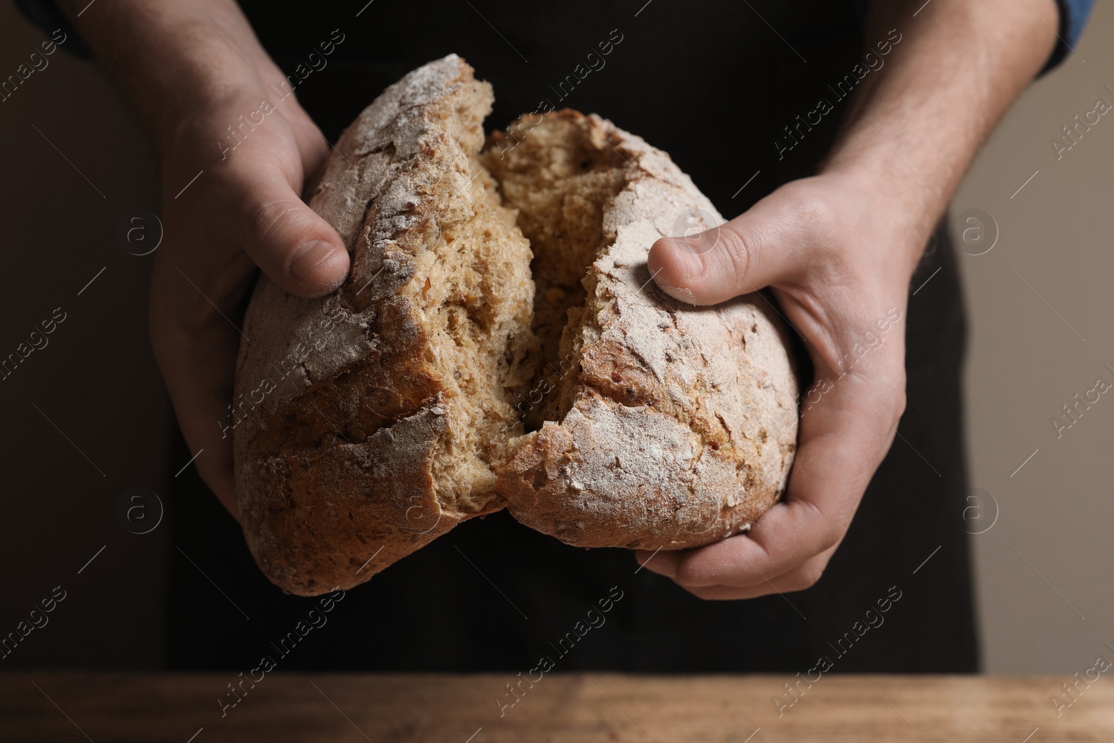 Photo of Man breaking loaf of fresh bread at wooden table, closeup