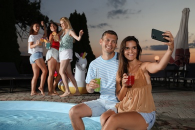 Happy couple with refreshing drinks taking selfie at pool party in evening