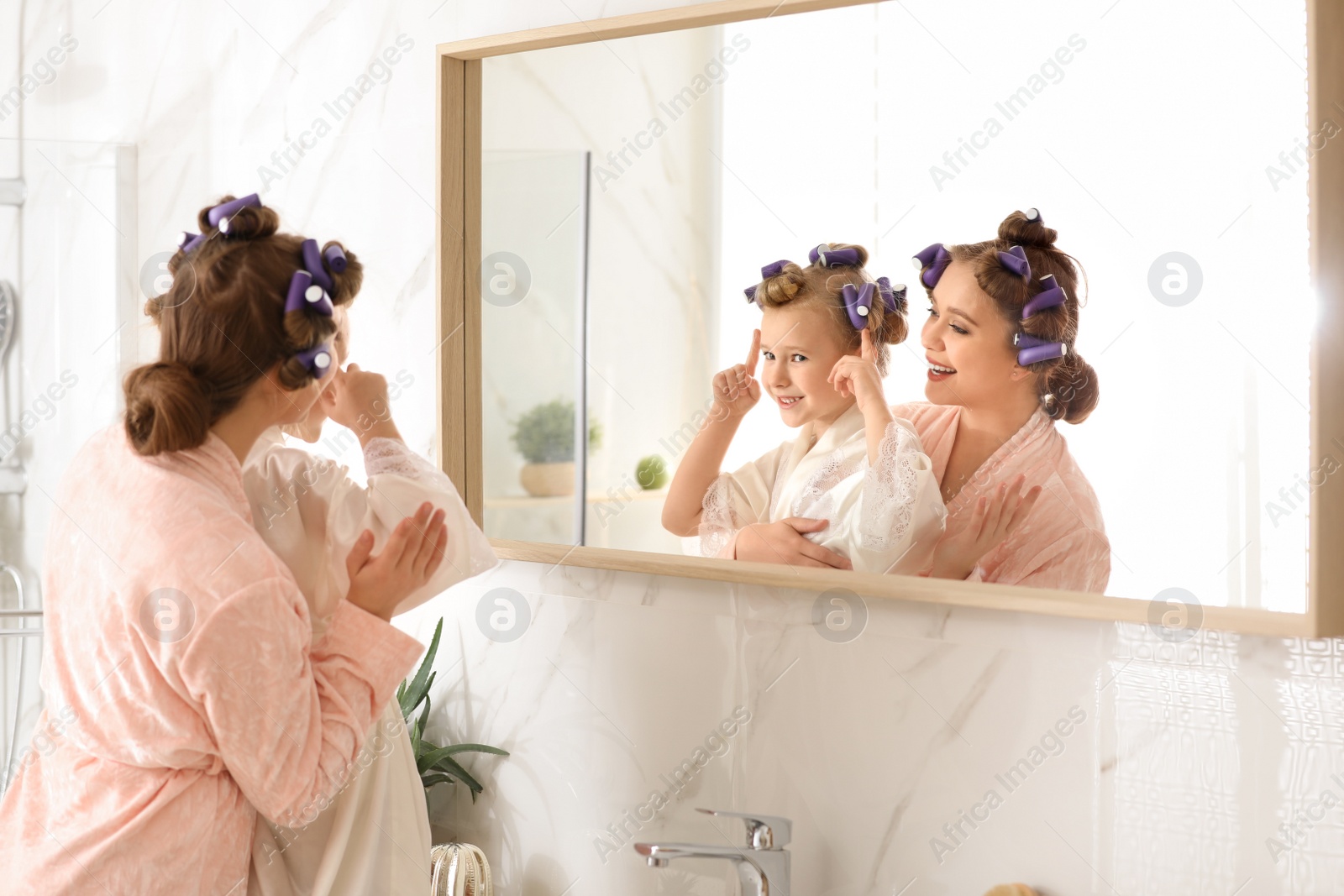 Photo of Happy mother and daughter with curlers near mirror in bathroom