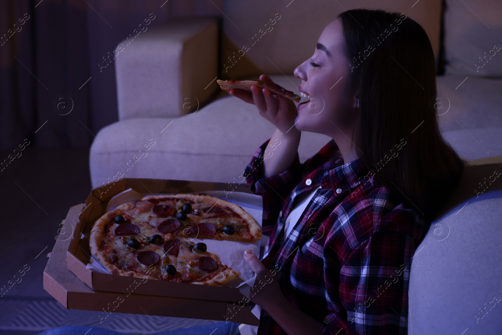 Photo of Young woman eating pizza while watching TV in room at night. Bad habit