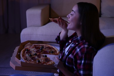 Photo of Young woman eating pizza while watching TV in room at night. Bad habit