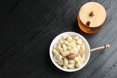 Peeled garlic cloves in bowl and honey on black table, flat lay. Space for text