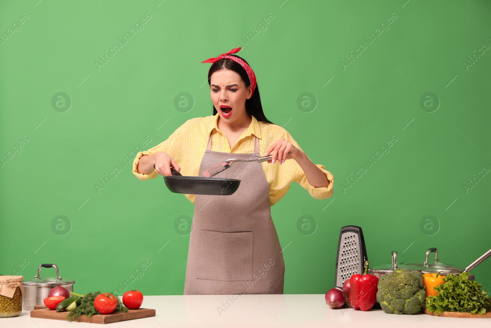 Photo of Emotional housewife with vegetables and different utensils on green background