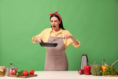 Photo of Emotional housewife with vegetables and different utensils on green background