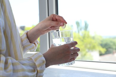 Photo of Woman putting effervescent pill into glass of water indoors, closeup