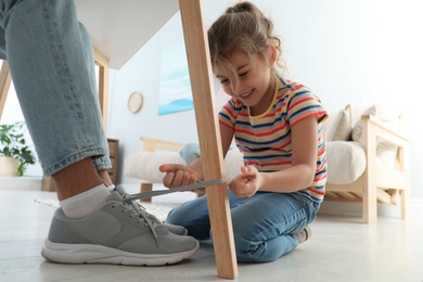 Photo of Cute little girl tying shoe laces of her father together at home, closeup