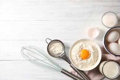 Photo of Flour with yolk in bowl and other ingredients for dough on white wooden table, flat lay. Space for text