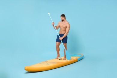 Handsome man with paddle on SUP board against light blue background