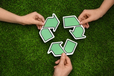 People holding paper recycling symbol above green grass, top view