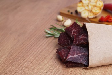 Photo of Paper bag with pieces of delicious beef jerky and different spices on wooden table, closeup. Space for text