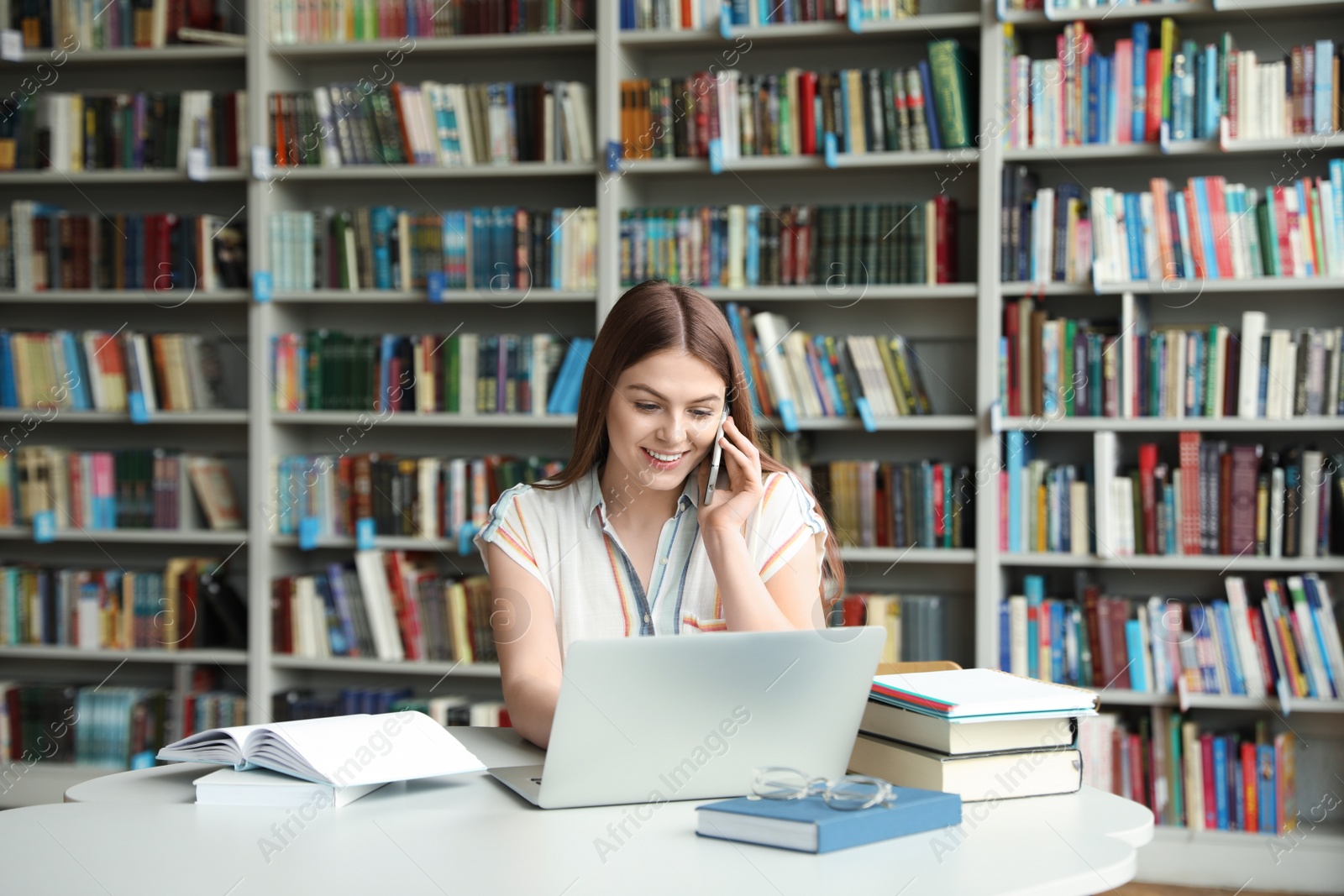 Photo of Young woman talking on phone and working with laptop at table in library