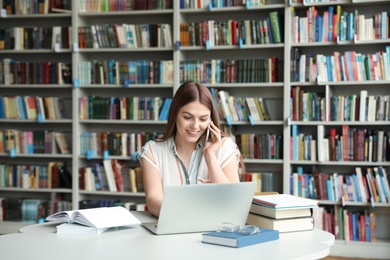 Photo of Young woman talking on phone and working with laptop at table in library