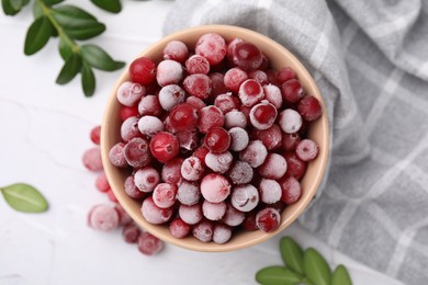 Frozen red cranberries in bowl and green leaves on white table, top view