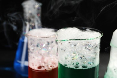 Laboratory glassware with colorful liquids and steam on black background, closeup. Chemical reaction