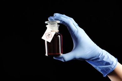 Photo of Woman in gloves holding glass bottle of poison with warning sign on black background, closeup