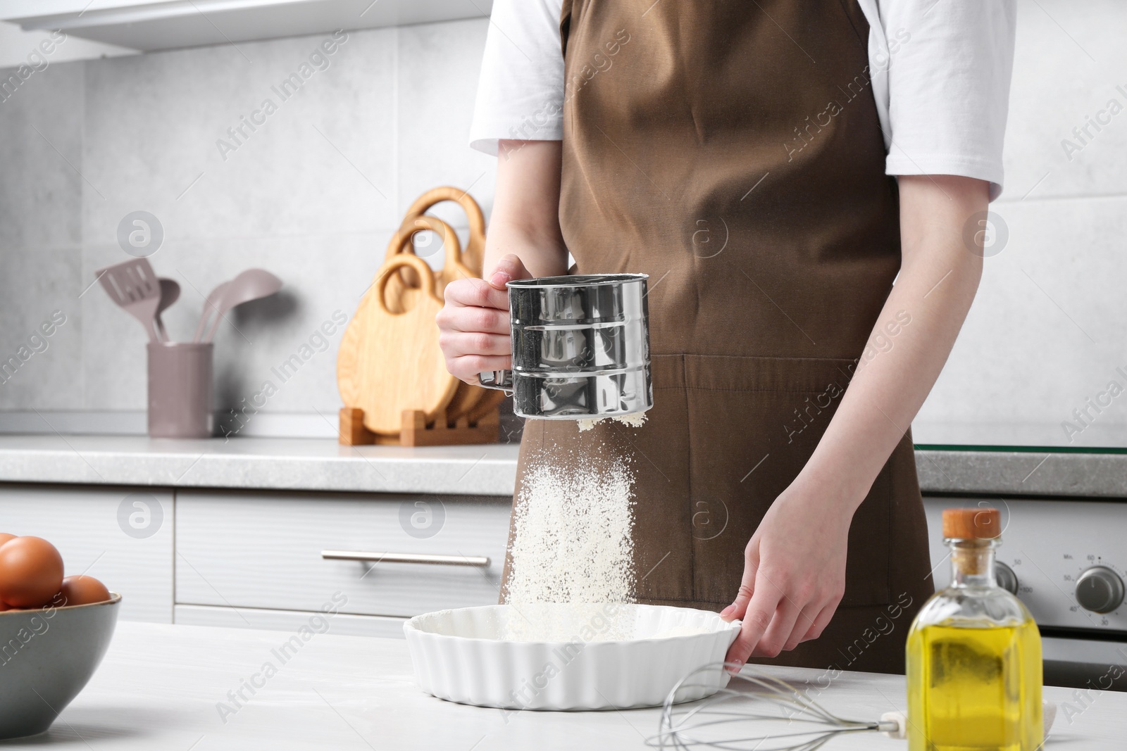 Photo of Woman sieving flour into baking dish at table in kitchen, closeup