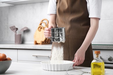 Photo of Woman sieving flour into baking dish at table in kitchen, closeup