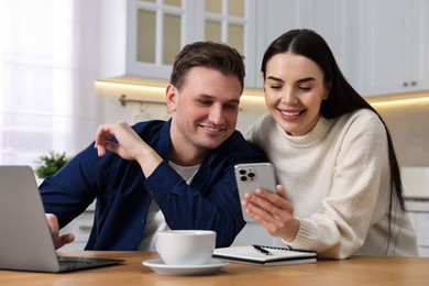 Photo of Happy couple using gadgets together at wooden table in kitchen
