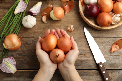 Photo of Woman holding yellow onion bulbs over wooden table, top view