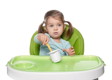 Photo of Cute little child eating tasty yogurt from plastic cup with spoon in high chair on white background