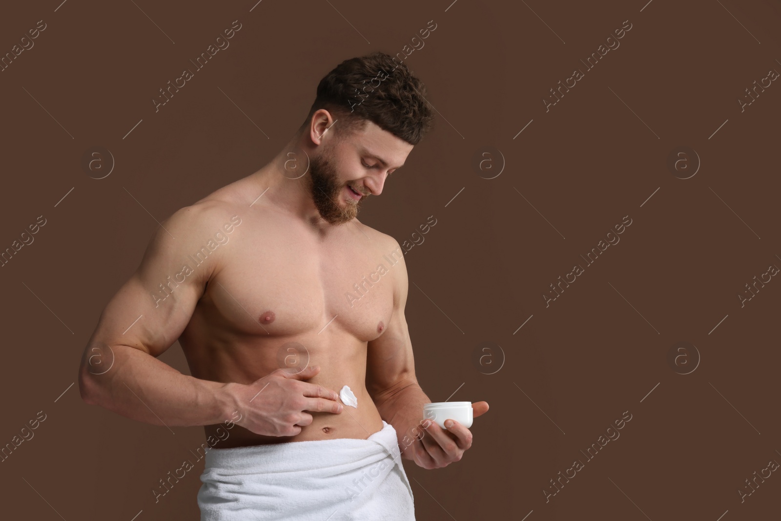 Photo of Handsome man applying moisturizing cream onto his body on brown background, space for text