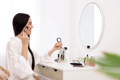 Photo of Beautiful young woman applying blusher with brush at dressing table indoors
