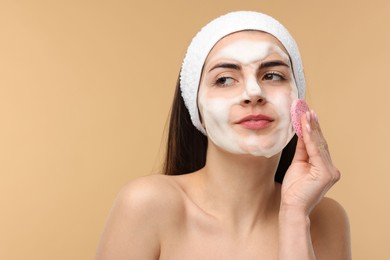 Photo of Young woman with headband washing her face using sponge on beige background, space for text
