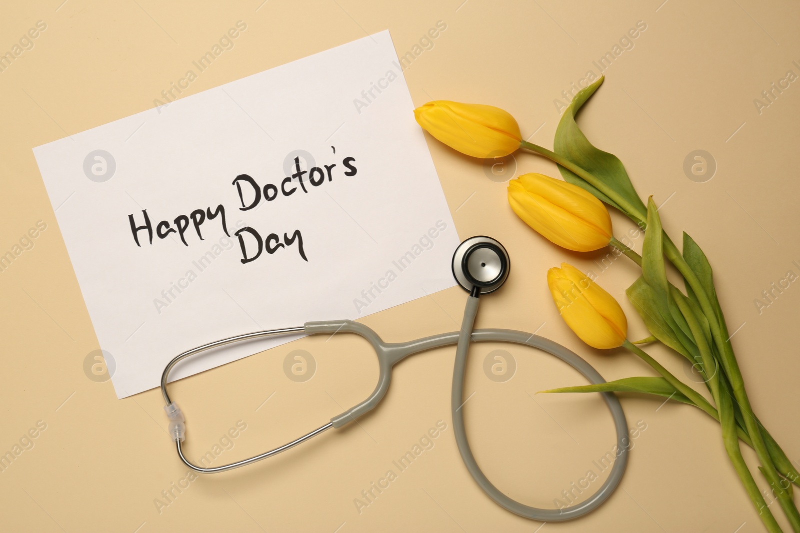 Photo of Card with phrase Happy Doctor's Day, stethoscope and yellow tulips on beige background, flat lay