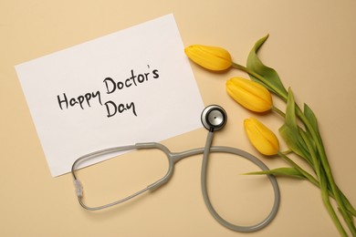 Card with phrase Happy Doctor's Day, stethoscope and yellow tulips on beige background, flat lay