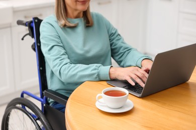 Photo of Woman in wheelchair using laptop at table indoors, closeup