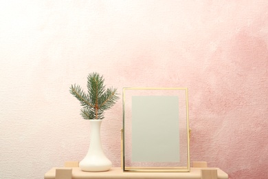 Photo of Blank frame and vase with fir branch on table near color wall. Mock up for design