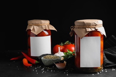 Photo of Jars of pickled tomatoes with blank stickers and products on black table