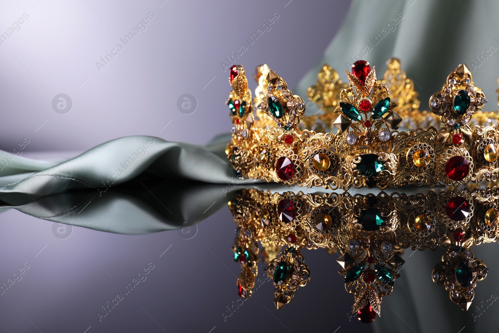 Photo of Beautiful golden crown with gems near light cloth on dark mirror surface. Space for text